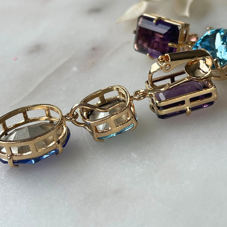 Ein Paar goldfarbene Clip-Ohrringe, ausgestattet mit austauschbaren Hängekristallen in Violett, Aquamarin- und Saphirblau, die für vielseitigen Glanz und Eleganz sorgen.